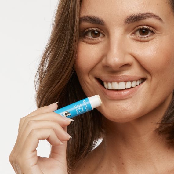 Close up of smiling female with olive skin and short brown hair applying the MooGoo Natural SPF15 Lip Balm. 