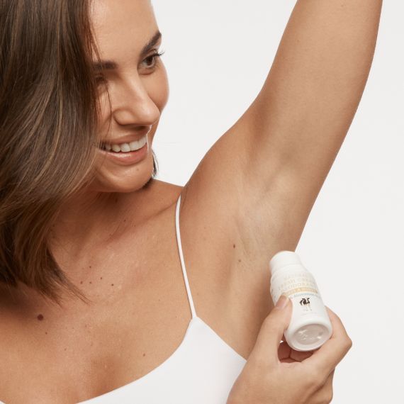 Brunette, olive skinned female model smiling and lifting her left arm to apply the MooGoo Fresh Cream Deodorant in Oats & Honey. 