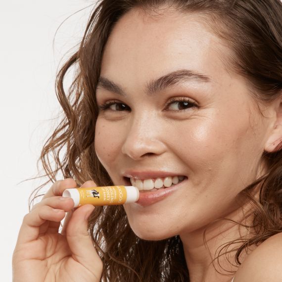 Smiling female model with curly brown hair applying the MooGoo Natural Tingling Honey lip Balm to her lips while looking at the camera. 