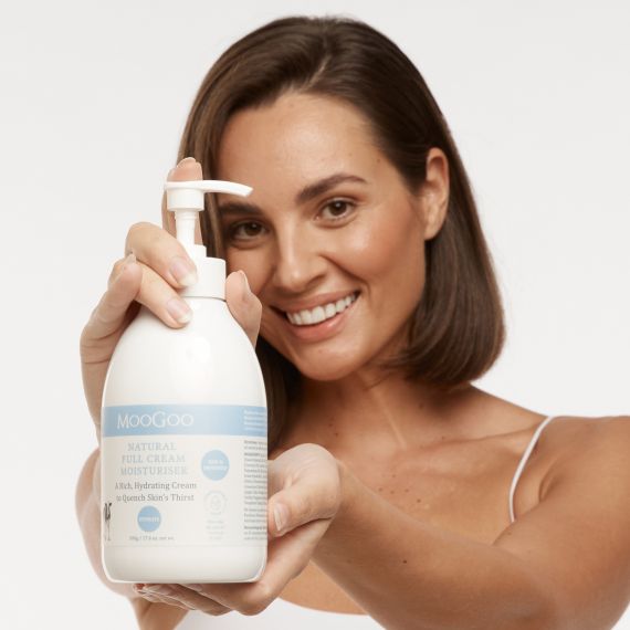 Smiling female model with short brown hair and olive skin holding the MooGoo Full Cream Moisturiser 500g bottle up to the camera with both hands. 