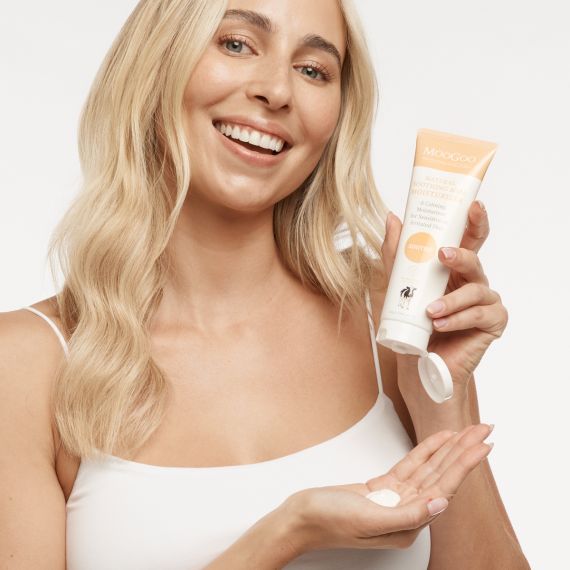 A blonde female model smiling at the camera while holding the MooGoo Natural Soothing MSM Moisturiser tube with her left hand and showing a squirt of the product in her right hand. 