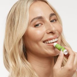 Smiling blonde haired, blue eyed female model applying the MooGoo Natural Cow Lick Lip Balm.