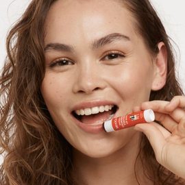Smiling female model with mixed islander and western heritage, brown curly hair and brown eyes applying the MooGoo Natural Strawberry Tinted Lip Balm. 