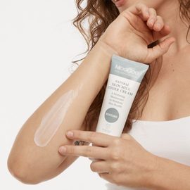 Female model with curly brown hair holding the MooGoo Natural Skin Milk Udder Cream next to a swatch of the product on her arm.